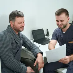 Lekarz w granatowym uniformie pokazuje pacjentowi w szarym szlafroku dokumenty podczas wizyty.