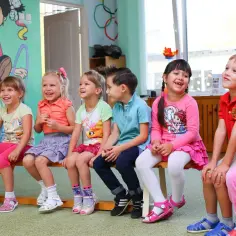 Grupa dzieci siedzi na ławce, uśmiechając się i patrząc w górę.