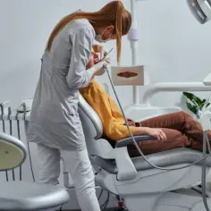 Dentystka w masce i rękawiczkach wykonuje zabieg pacjentowi siedzącemu na fotelu dentystycznym.